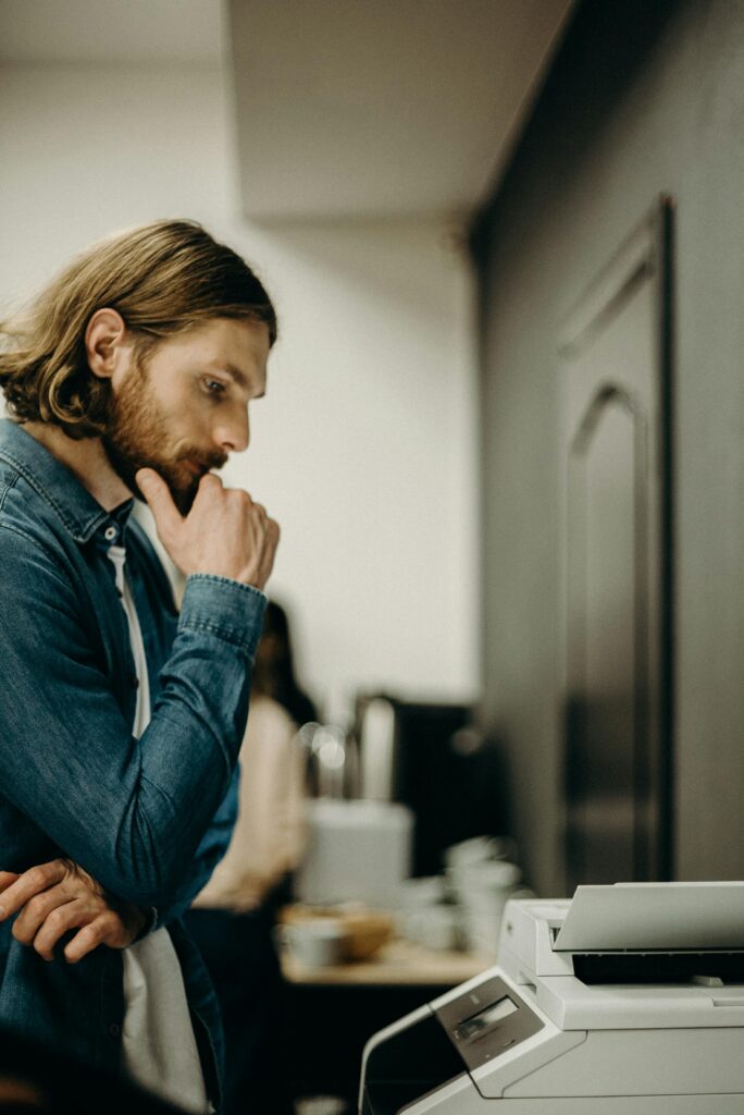 A man in deep thought while operating a printer in an office setting.